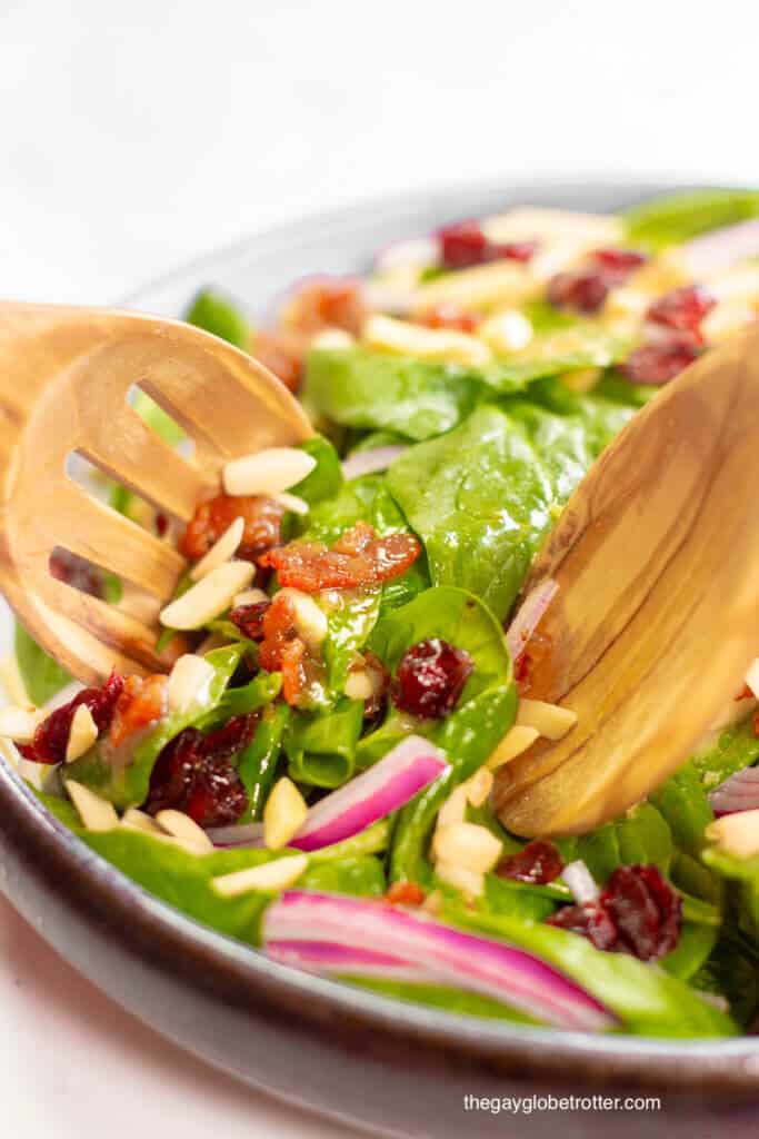 Serving utensils tossing spinach cranberry salad and warm bacon dressing.