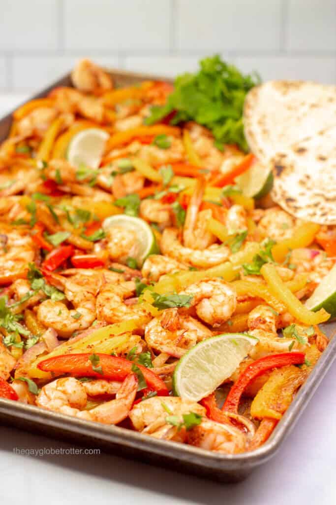 Shrimp fajitas being served on a baking sheet with cilantro, limes, and tortillas.