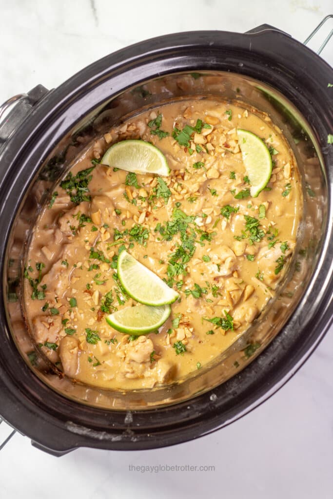 A slow cooker filled with thai peanut chicken garnished with limes, cilantro, and peanuts.