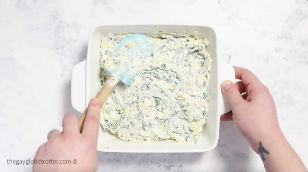 Spinach artichoke dip being spread into a baking dish with a spatula.