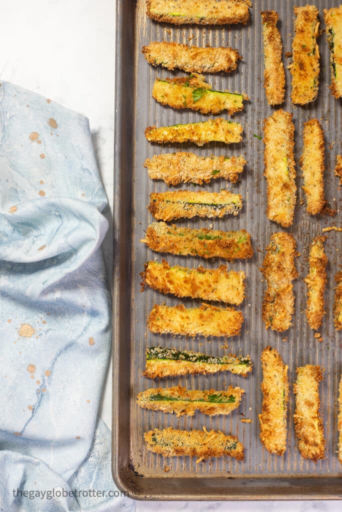 A baking sheet with baked zucchini sticks next to a tea towel.